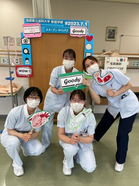 看護学部の学生たちがオープンキャンパスで楽しそうにポーズをとり、手に持ったサインボードには「Goody」や医療関連のイラストが描かれています。