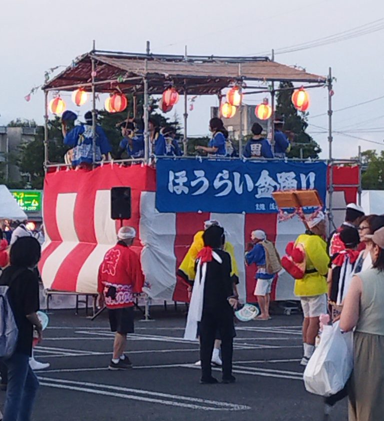 夏祭りの櫓の上で踊る人々と、周囲の参加者が賑わう様子。