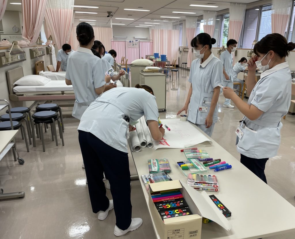 看護学部のオープンキャンパス準備中、学生たちが「血圧測定コーナー」のポスターを作成している様子。