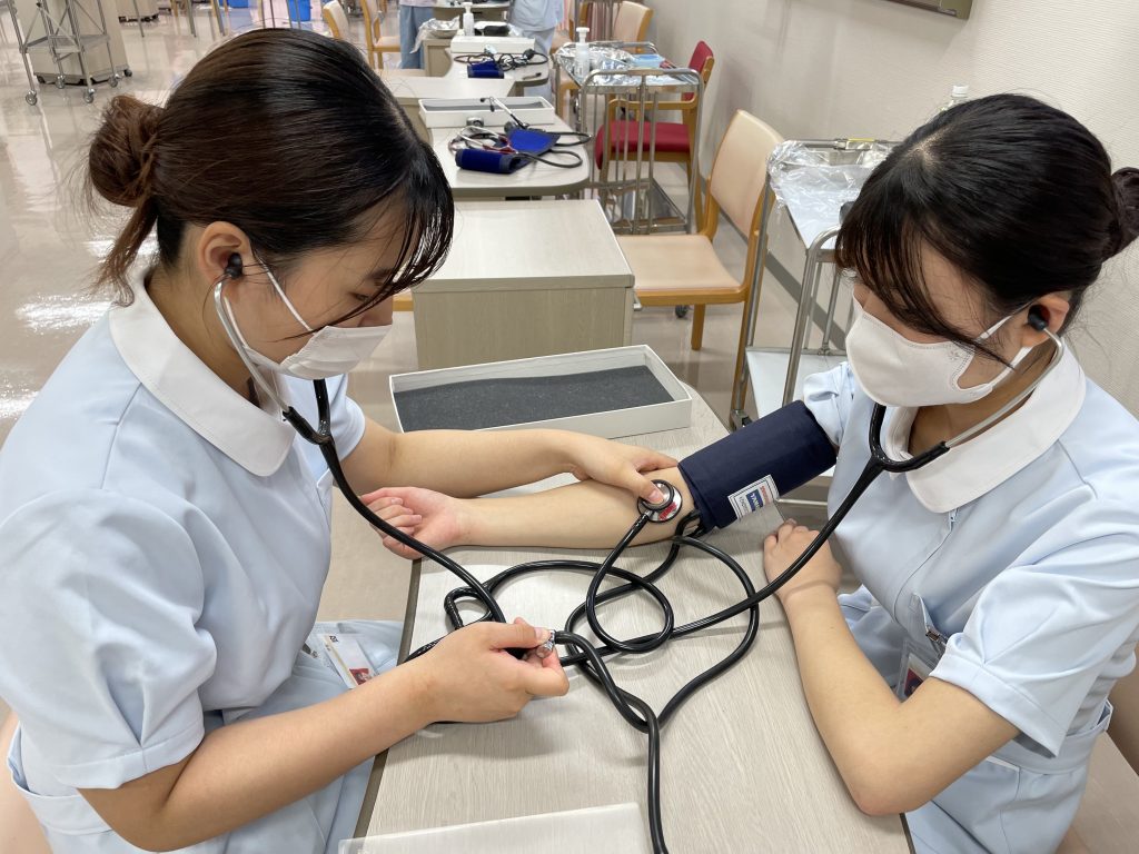 看護学部のオープンキャンパスで、学生が血圧測定の練習をしている様子。マンシェットの巻き方が上手です。