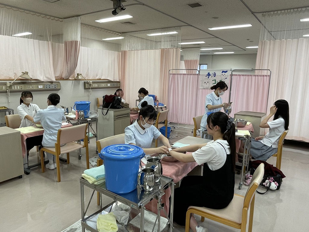 看護学部のオープンキャンパスで手浴コーナーを体験する学生たちの様子。リラックスした雰囲気が漂っています。