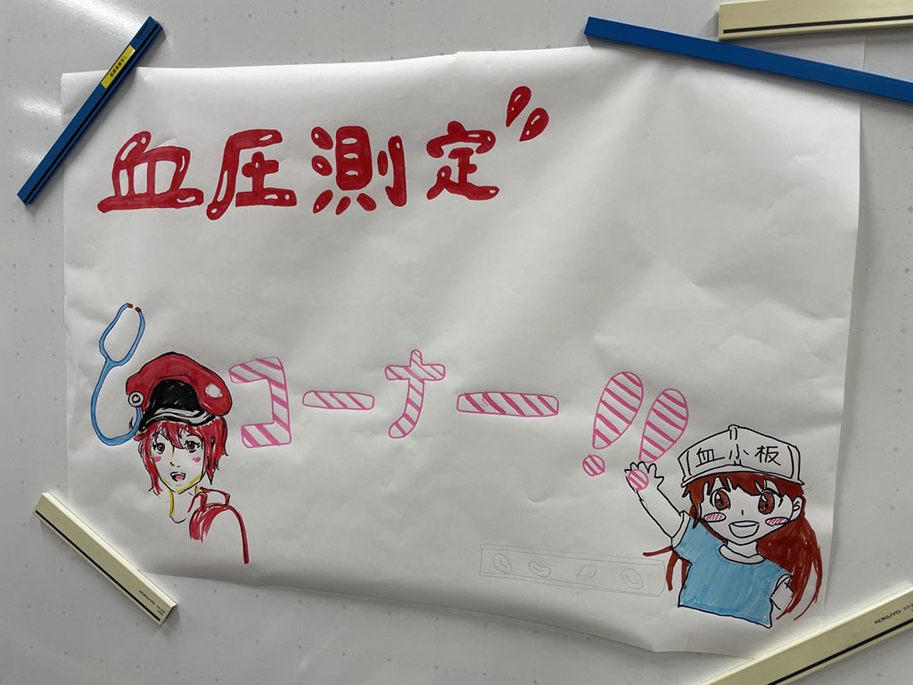 血圧測定のコーナーを紹介するカラフルなポスター、看護学部の学生が描いたイラスト付き。