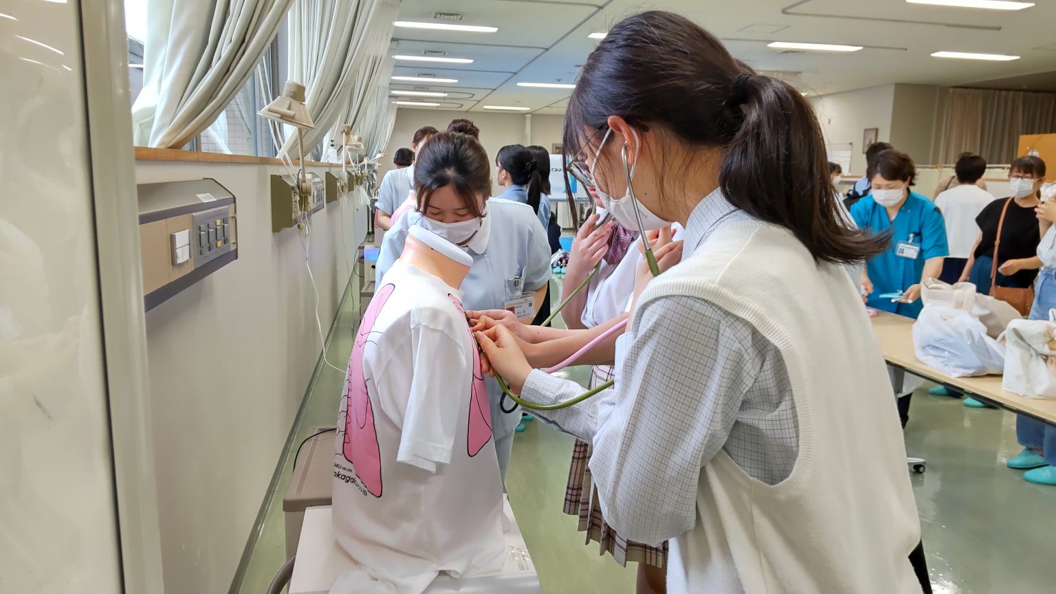 看護学部のオープンキャンパスで、学生が模擬患者に聴診器を使っている様子。