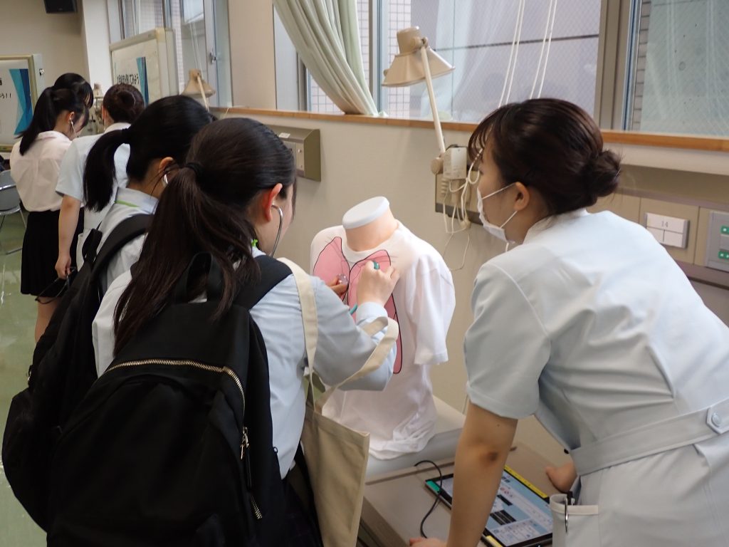 看護学部オープンキャンパスで、学生が実習用の模型に触れながら教員と交流している様子。
