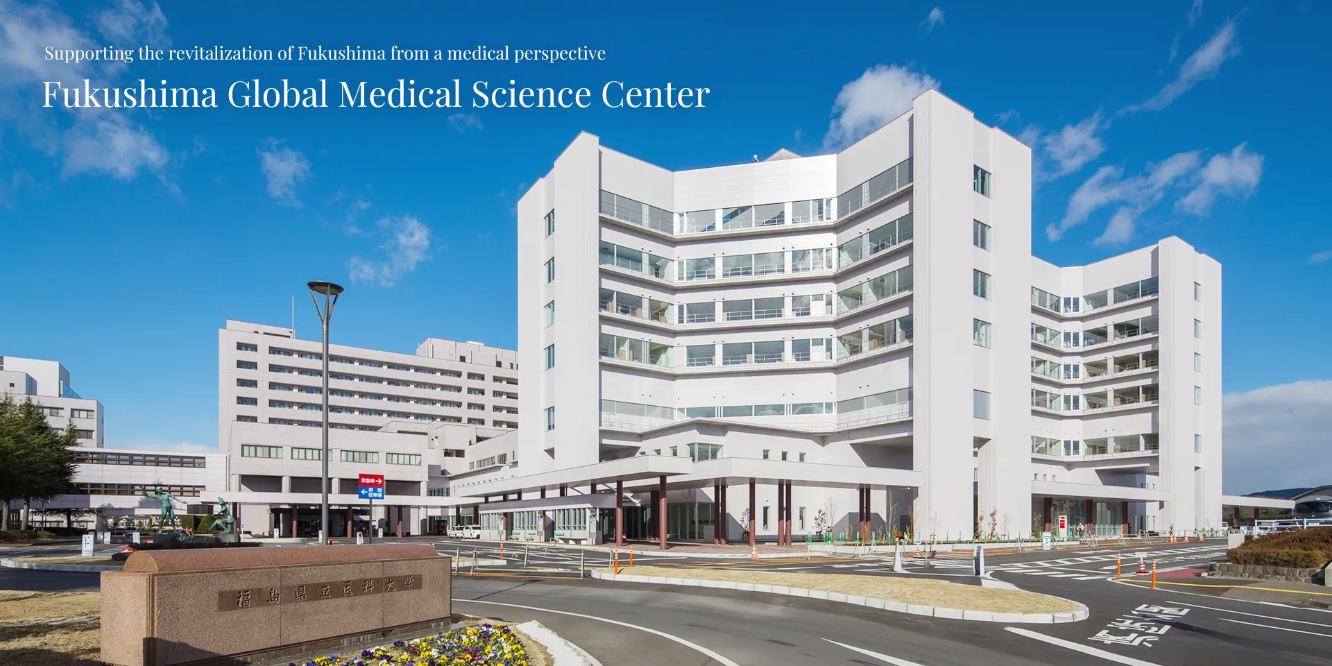 A photograph capturing the exterior of the Fukushima Global Medical Science Center and its surrounding landscape, featuring modern architecture under a clear blue sky.