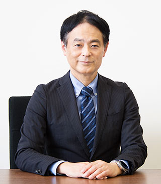 A man in a dark suit sits at a desk with his hands folded.