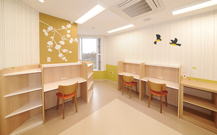 プレイルームの学習スペースで、木製の机と椅子が配置され、壁には可愛らしい鳥のイラストが描かれています。