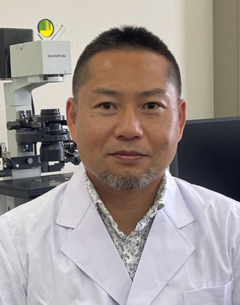 井上直和教授が白衣を着て微生物学の研究室で微細な機器の前に座っている写真。