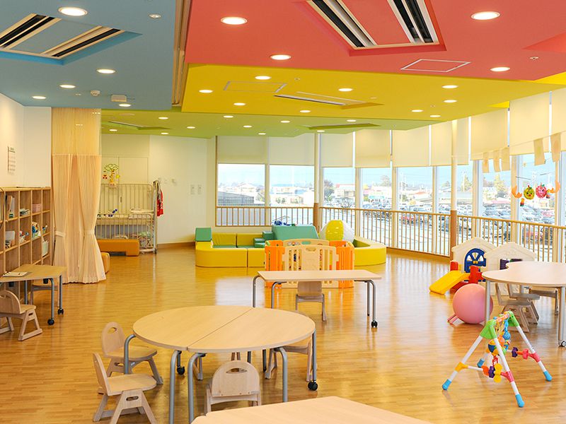 明るい色の天井と木製の床が特徴の保育室。テーブルや椅子、遊具が配置され、外は大きな窓からの景色が見える。