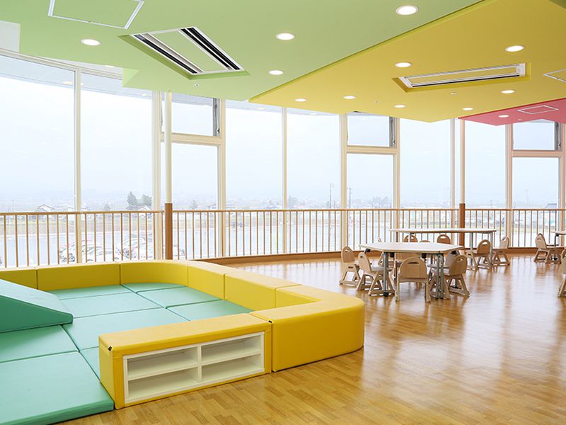 広々とした明るい室内に、窓に面したテーブルと椅子、柔らかい素材の囲いスペースがあり、天井にはカラフルなパネルが設置されています。