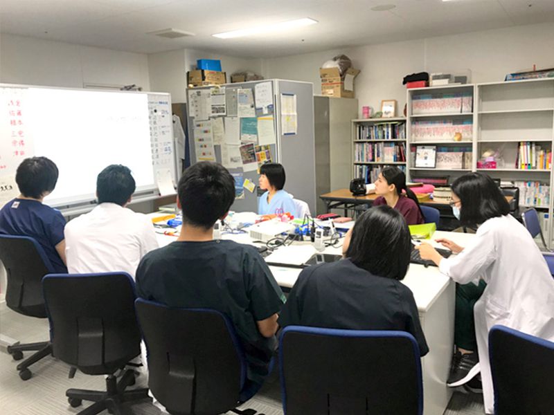 大学の教室で医学生たちがホワイトボードの前に座り講義を受けています。背景には本棚が見えます。