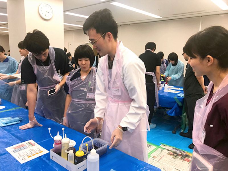 医学生が実習で作業を行う様子。白衣やエプロンを着用し、机には色付きの液体の入ったボトルが並んでいる。