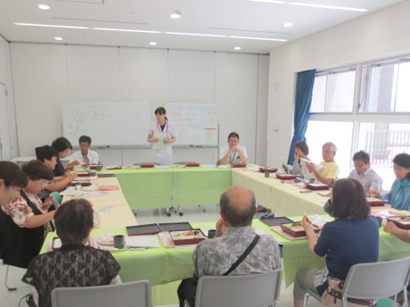 医療従事者が会議室で対面し、真ん中に立つ人物が説明を行っている。テーブルには食事が並んでいる。