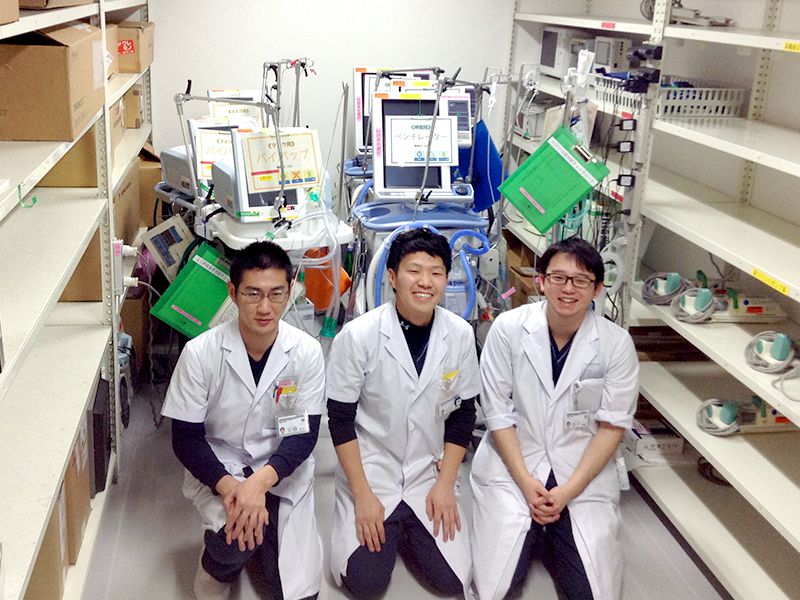 白衣を着た三人の医学生が病院の備品室で機器の前に座って微笑んでいる。周囲には棚が並んでいる。