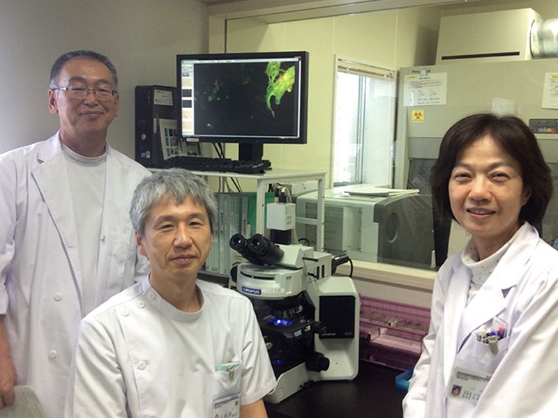 研究室で白衣を着た三人の医療スタッフが顕微鏡を前に微笑んでいる。背景には細胞の画像が表示されたモニターがある。