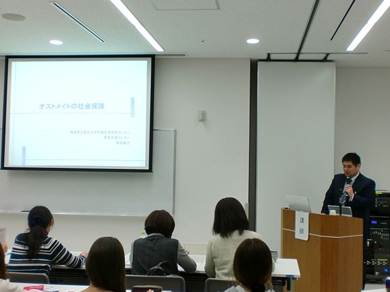 教室で講師が講演をしている様子。プロジェクター画面に「オストメイトの社会保障」と表示され、数名の聴衆が座って聞いている。