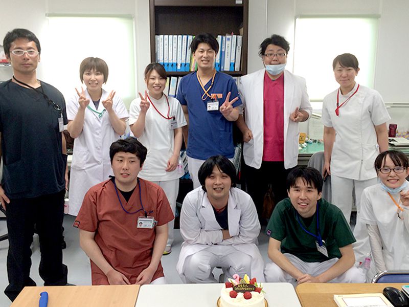 お祝いのケーキを前に、笑顔でポーズをとる医療従事者のグループ。白衣やスクラブを着用し、後方には書類が整理された棚が見える。