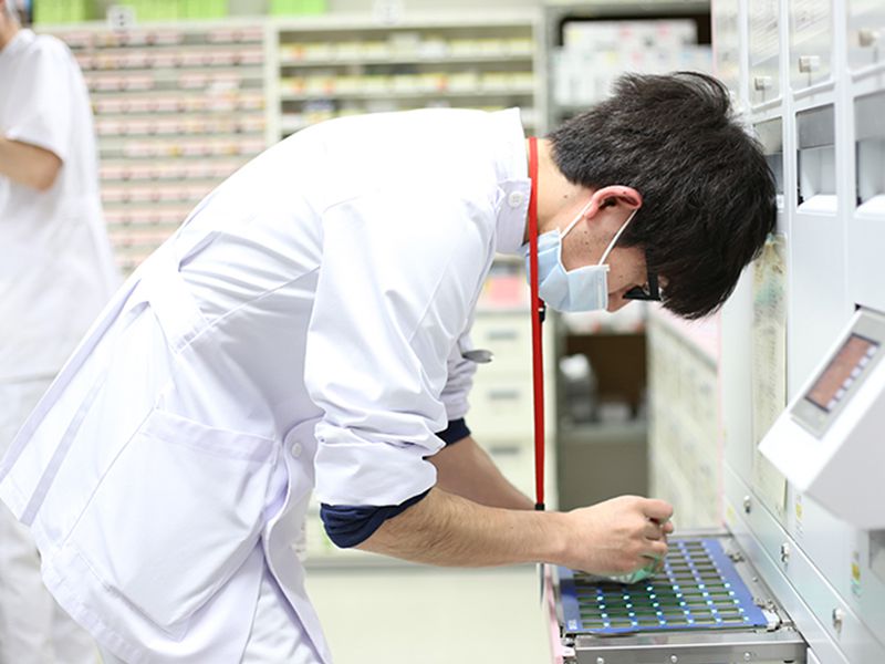薬局内の作業台で、マスクを着けた白衣の医療従事者が細かい作業をしている。背景には薬棚が並ぶ。