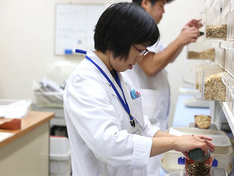 研究室で作業する白衣を着た二人の医療関係者が、棚にある複数のトレイから材料を計量しています。