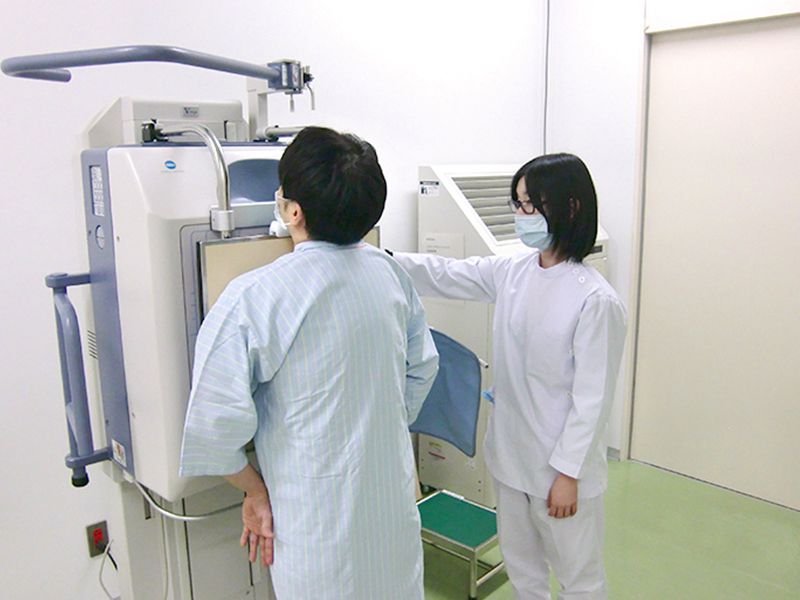 患者が病院でマスクを着けて検査を受けている様子。医療スタッフが横でサポートしています。白い壁と医療機器が背景にあります。