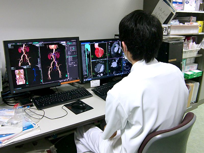 医療従事者が2台のモニターで心臓や血管の画像を解析している場面。机上に医療関連資料が散在。