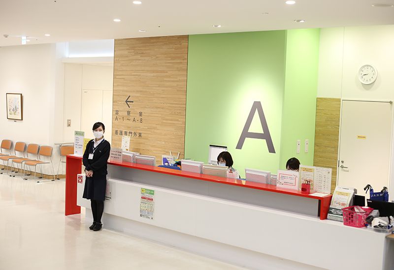 病院の受付カウンター前に立つマスクをした女性スタッフと、カウンターで作業する別のスタッフ。