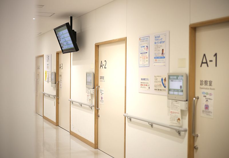 白い壁に並ぶ診察室のドアがあり、それぞれ案内板等が設置された病院の廊下。