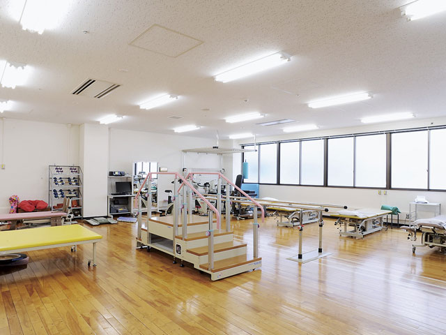広々としたリハビリテーション室で、木製の床や明るい照明の下にリハビリ機器とトレーニングスペースが整然と配置されている。