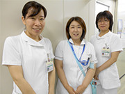 3人の看護師が制服を着て廊下で笑顔を見せている。彼女たちは医療機関で働いているように見える。