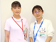 二人の女性が並んで立っており、白衣やIDバッジを身に着けている。背景はシンプルな白壁。