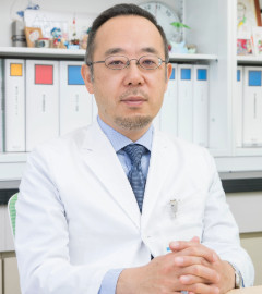 腫瘍内科の主任教授、佐治重衡氏が白衣を着て座っている写真。背景にはファイルが並んでいる。
