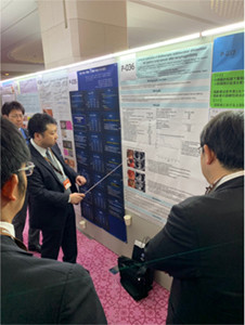 会場にて研究内容をパネル前で解説中の様子。