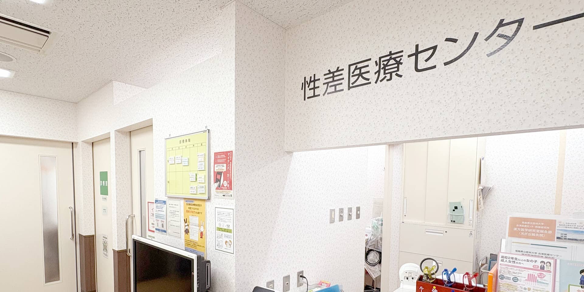 医療施設の受付エリアで、壁に「性差医療センター」と書かれた標識がある。診察室のドアとパンフレットが見える。