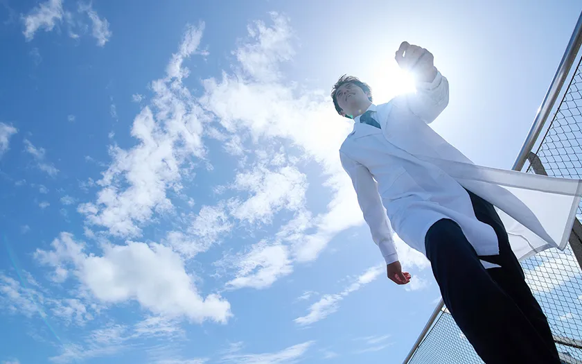 医療白衣を着た男性が青空の下で歩いている様子。背景には明るい太陽と雲が広がっている。