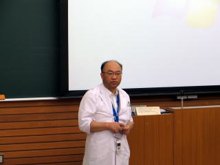 大谷晃司先生が教室で講義をしている様子。白衣を着た先生が前に立ち、背後にはスクリーンと木目調の壁がある。