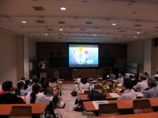 講堂でプレゼンテーションを聴く大学生たち。スクリーンにカラフルな図が映し出されている。