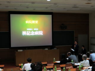 講義室でスライドを使った発表が行われており、聴衆が座っている場面。スライドには「病院概要」と書かれている。