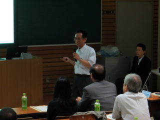 講堂で講演中の男性がプロジェクターを使って説明しており、数名の聴衆が机に座って聞いている。