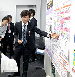 スーツ姿の男性がカンファレンスでポスター発表を行い、指差しながら説明している。後ろに数人の見学者がいる。