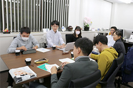 会議室で、マスクをした数人の男女がテーブルを囲んで資料を見ながら議論している様子。背景には窓がある。