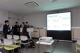 教室で学生たちがスクリーンに表示されたグラフを使ってプレゼンテーションをしている様子。前に座る他の参加者たちも注目している。