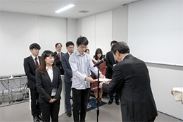 大学の授賞式で、スーツ姿の学生が証書を受け取っている様子。他の学生も列に並び見守っている。