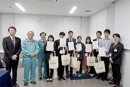 数名の大学生や関係者が賞状とバッグを持ち、室内で記念撮影をしている。背景には白い壁がある。