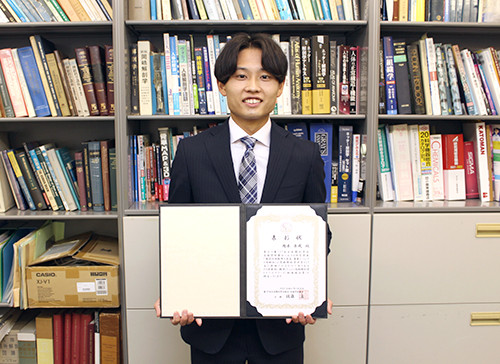 スーツを着た男性が賞状を手に持ち、本棚の前で微笑んでいる。様々な書籍が背景に並ぶ。