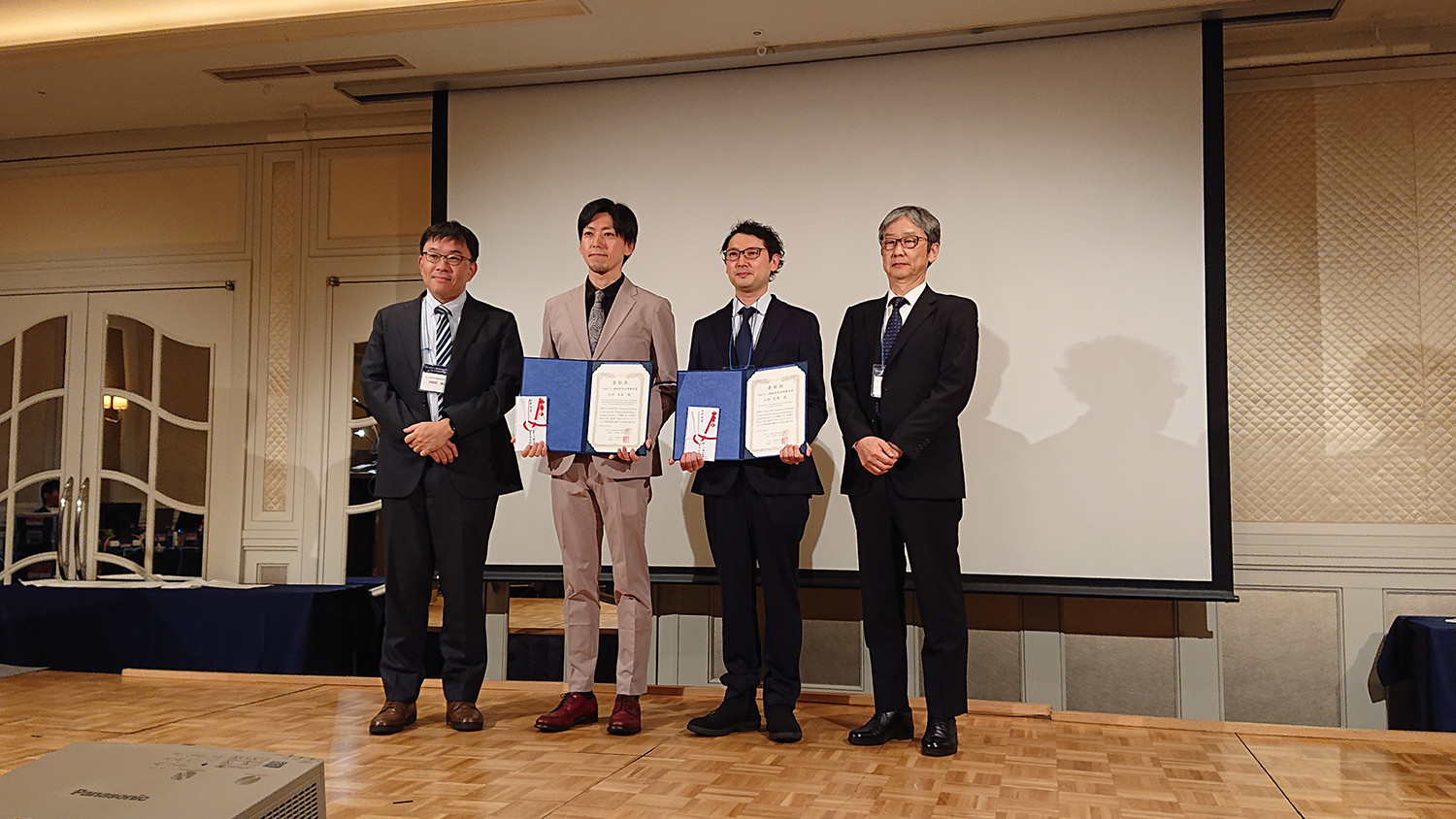 4人の男性が表彰式で賞状を持ち、スーツを着て舞台上に立っている。背景にスクリーンと装飾的な壁がある。