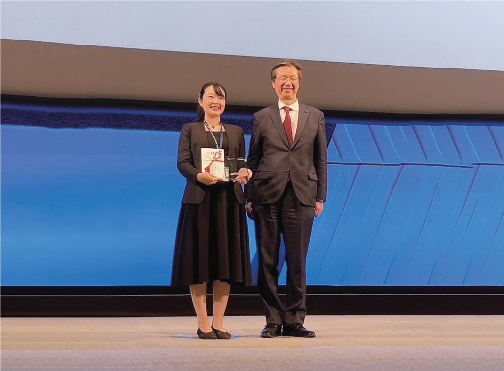 壇上で賞を手にする二人の人物。背景は青を基調とした装飾が施されている。