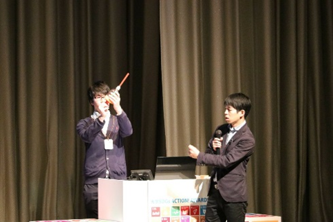 2人の男性がステージ上でマイクを使いプレゼンをしている。片方の男性は小道具を持ち説明している様子。