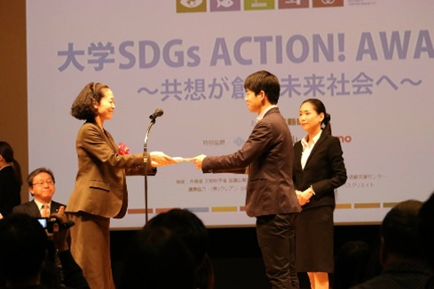 大学SDGsアクションアワードの授賞式で、壇上の人物がマイク前で賞を手渡しているシーン。観客が見守る。
