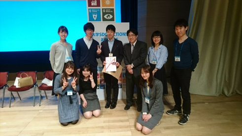 学生と大人がステージ上でポスターを持ち集まっている。背景にはSDGs関連のディスプレイが見える。