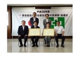 5人の大人が式典で表彰状を持って前列に座り、他の2人が後列で立っている。背景には表彰式の幕が掲示されている。